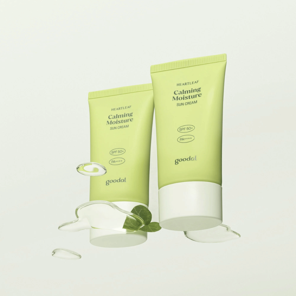 Twee tubes van Goodal Heartleaf Calming Moisture Sun Cream SPF50+ PA++++ omgeven door plantbladeren en serumdruppels tegen een lichte achtergrond.