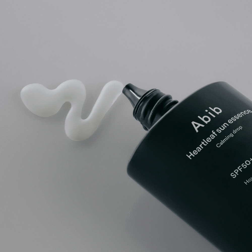 Close-up van Abib Heartleaf Sun Essence Calming Drop met de zonnebrandcrème uitgeperst in een witte, golvende lijn op een lichte ondergrond.