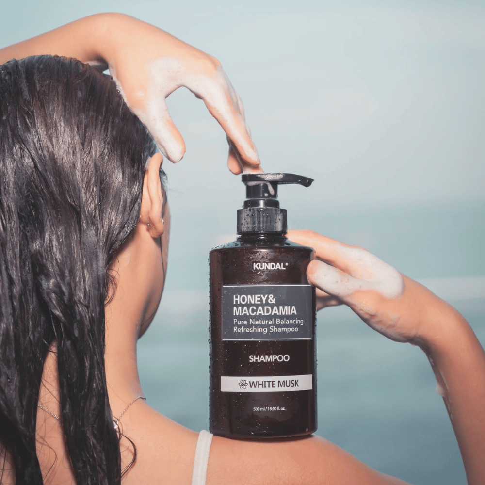 Vrouw met nat haar brengt schuim aan en houdt KUNDAL Honey & Macadamia Nature Shampoo White Musk tegen een achtergrond van de zee.