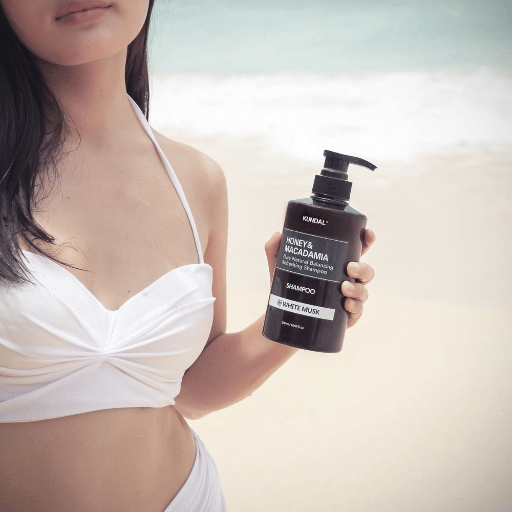 Vrouw in witte bikini houdt KUNDAL Honey & Macadamia Nature Shampoo White Musk op een strand met de zee op de achtergrond.