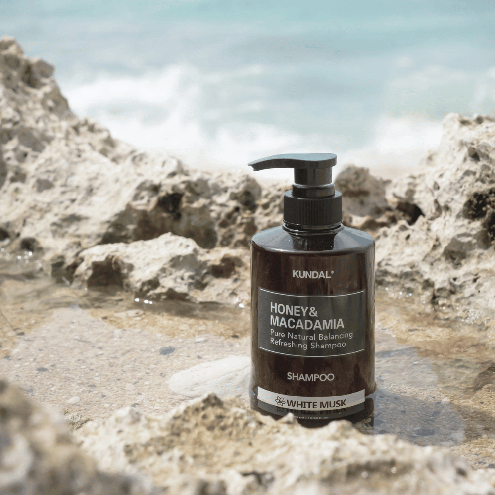 KUNDAL Honey & Macadamia Nature Shampoo White Musk geplaatst op rotsen bij de zee, een natuurlijke haarverzorgingsproduct met een frisse geur.