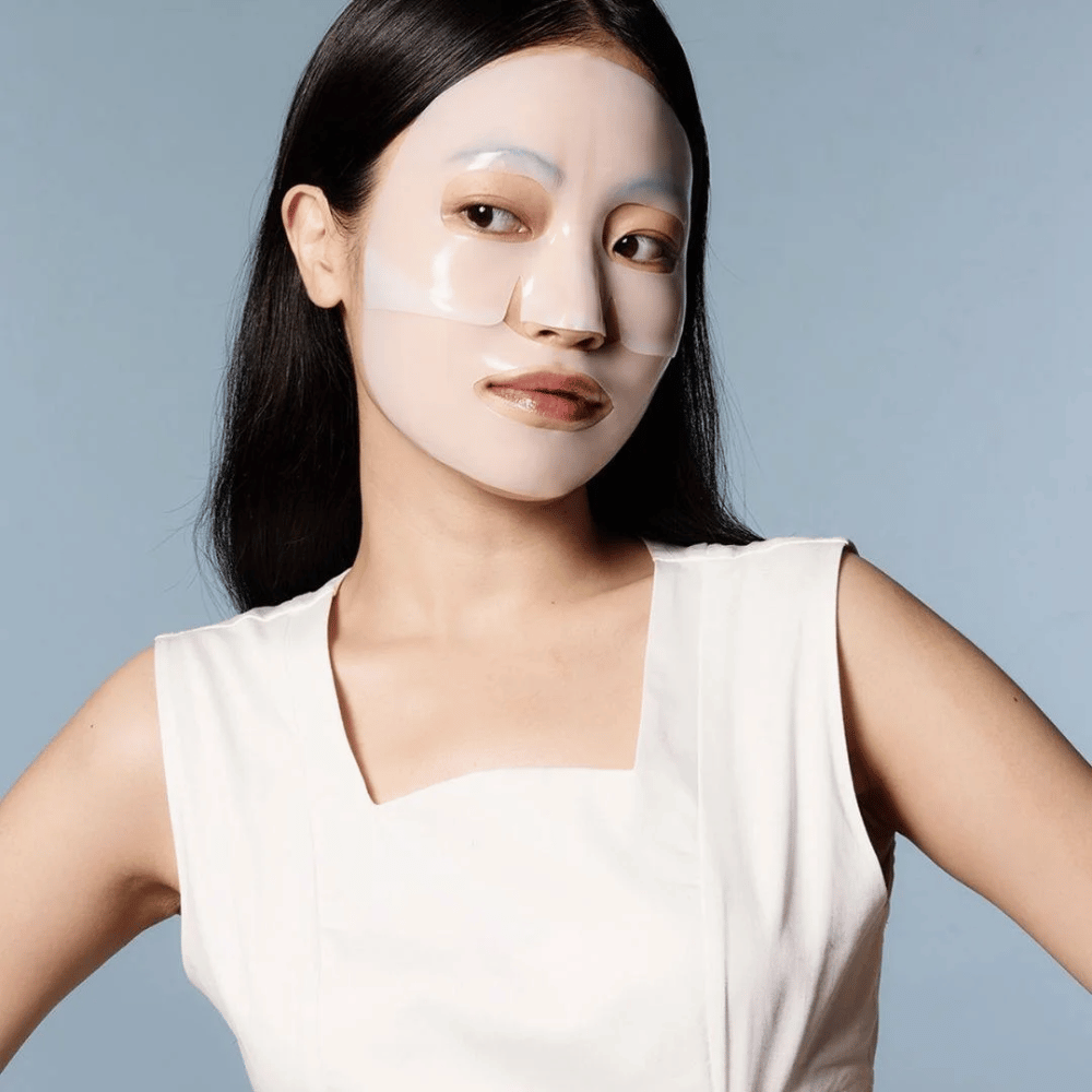 Een vrouw in een witte mouwloze jurk draagt een doorzichtig gezichtsmasker dat bijna haar hele gezicht bedekt. Het masker geeft de indruk van geavanceerde huidverzorging en heeft het uiterlijk van een schoonheidsbehandeling, ontworpen om de huid diep te hydrateren en te behandelen.
