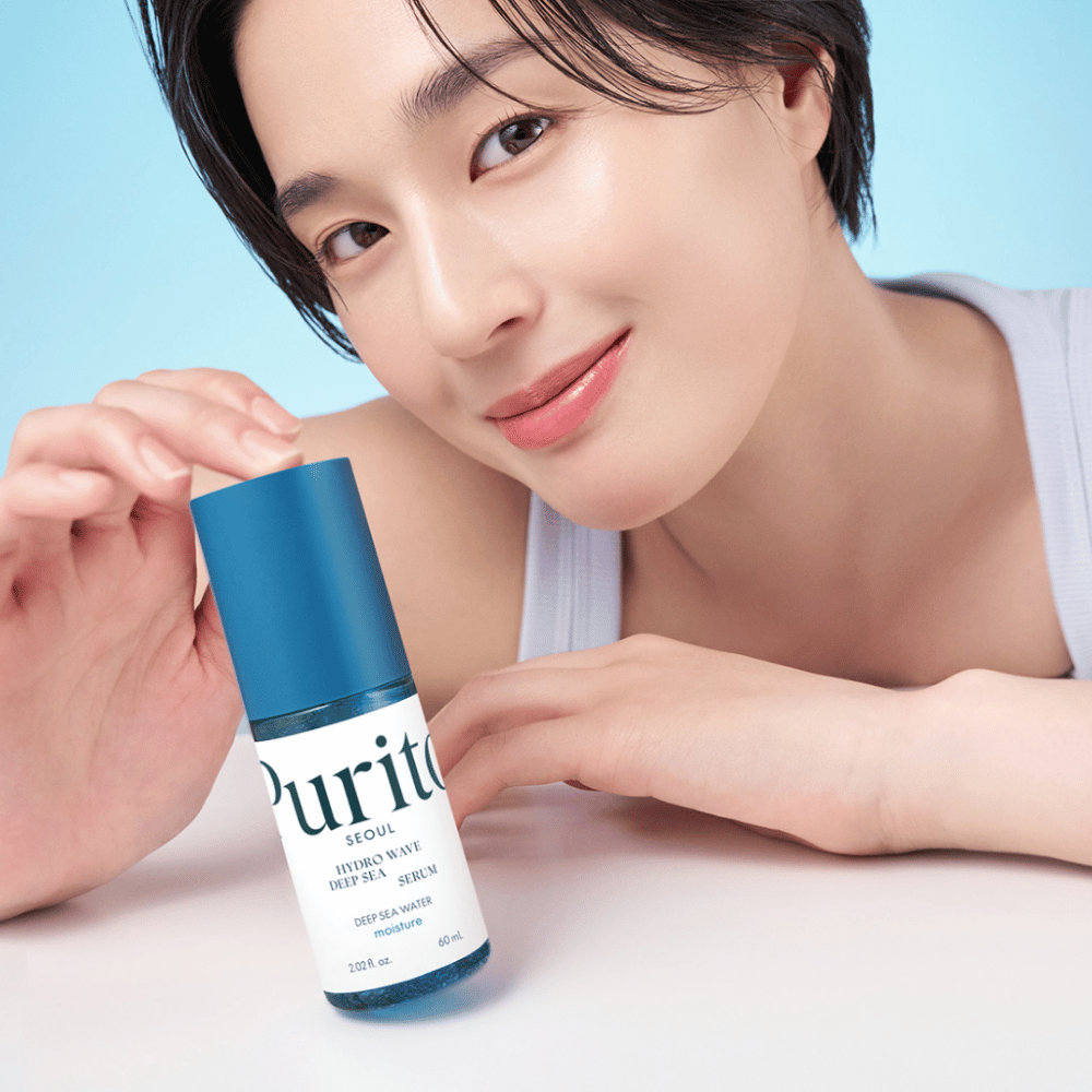 Een vrouw met kort haar houdt een blauw serum van Purito, Hydro Wave Deep Sea Serum. Haar huid ziet er stralend en vochtig uit, wat het product in de schijnwerpers zet. De achtergrond is lichtblauw, wat een frisse en schone uitstraling geeft. Het product is verdiept in huidverzorging.