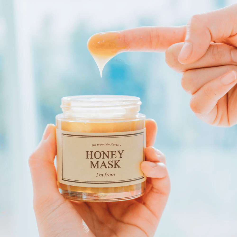 Een hand houdt een open pot "I'm from Honey Mask", en een vinger wordt in het gouden masker gedoopt tegen een lichte achtergrond.