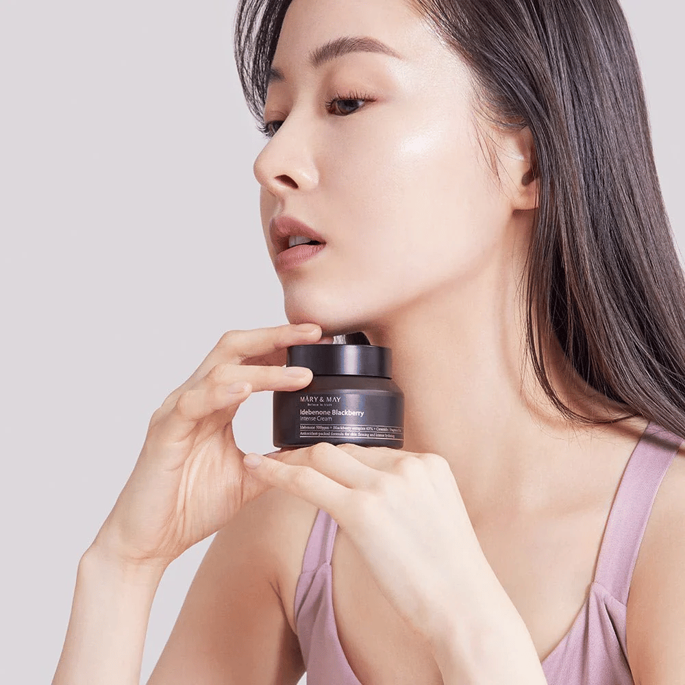 Een vrouw houdt een pot met MARY & MAY Idebenone Blackberry Intense Cream. Ze heeft een stralende huid en glimlacht zachtjes, gekleed in een mouwloze top. Het product is een intensieve crème met idebenon en bramencomplex bedoeld voor de stevigheid en hydratatie van de huid.