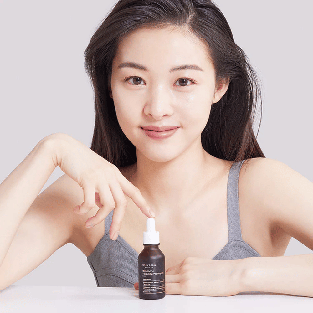 Een vrouw met lang, donker haar houdt MARY & MAY Idebenone + Blackberry Complex Serum vast, een donkere glazen fles met pipet. Ze heeft een brede glimlach en wijst naar de fles met haar andere hand. De vrouw draagt een grijze, mouwloze top en lijkt zich in een lichte en eenvoudige omgeving te bevinden, wat de focus van het product op huidverzorging en schoonheid benadrukt.