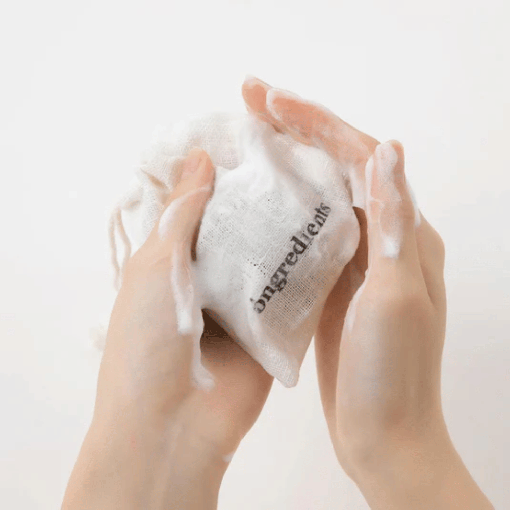 Handen die Óngredients Jeju Green Tea Cleansing Ball opschuimen in een stoffen zak – veganistische gezichtsreiniging met groene thee.