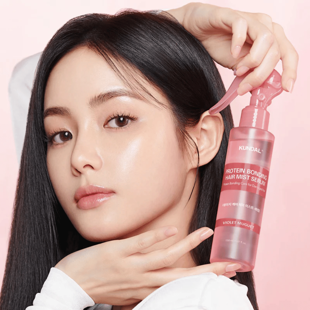 Vrouw met lang zwart haar houdt KUNDAL Protein Bonding Hair Mist Serum Violet Muguet voor haar gezicht, tegen een lichtroze achtergrond.