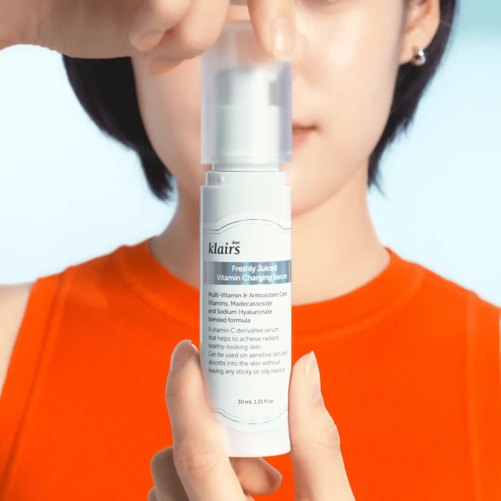 Vrouw in oranje trui houdt Klairs Freshly Juiced Vitamin Charging Serum voor zich tegen een blauwe achtergrond.