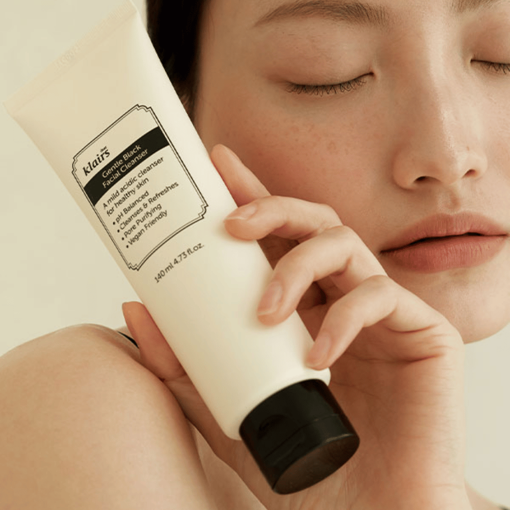 Een persoon houdt een tube van Klairs Gentle Black Facial Cleanser tegen zijn wang, gesloten ogen en natuurlijke huidskleur zijn zichtbaar op de achtergrond.