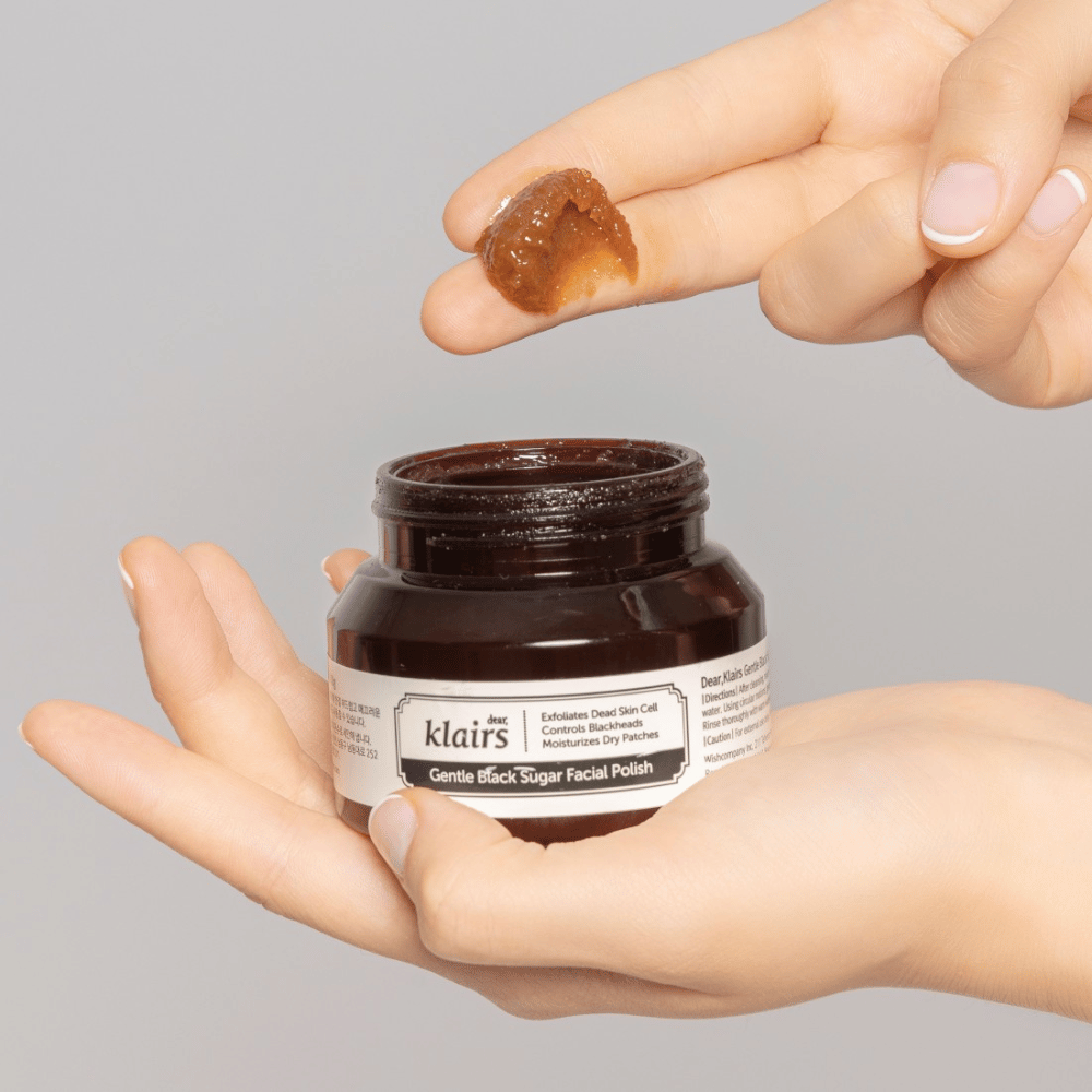 Handen houden een open pot van Klairs Gentle Black Sugar Facial Polish en tonen het product op een vinger, tegen een grijze achtergrond.