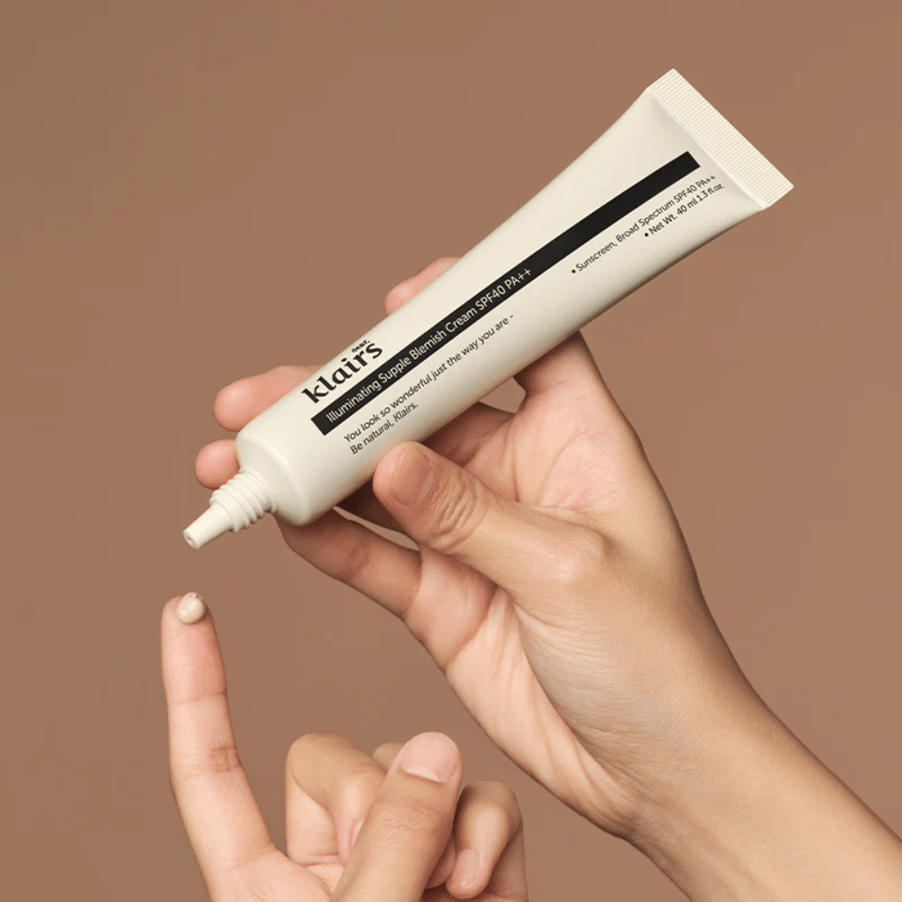 Een hand houdt een tube van Klairs Illuminating Supple Blemish Cream SPF40 PA++ en knijpt een kleine hoeveelheid crème op een vinger, tegen een bruine achtergrond.