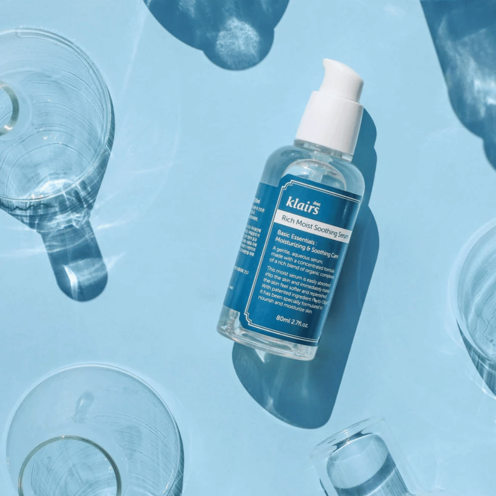 Een fles Klairs Rich Moist Soothing Serum tussen waterglazen met reflecties op een blauwe achtergrond.