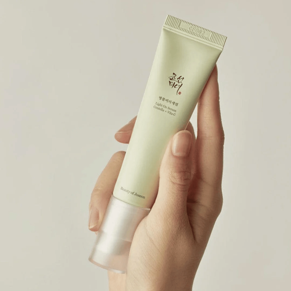 Een hand houdt Beauty of Joseon Light On Serum met Centella en vitamine C, in een lichtgroene tube met minimalistisch ontwerp tegen een neutrale achtergrond.