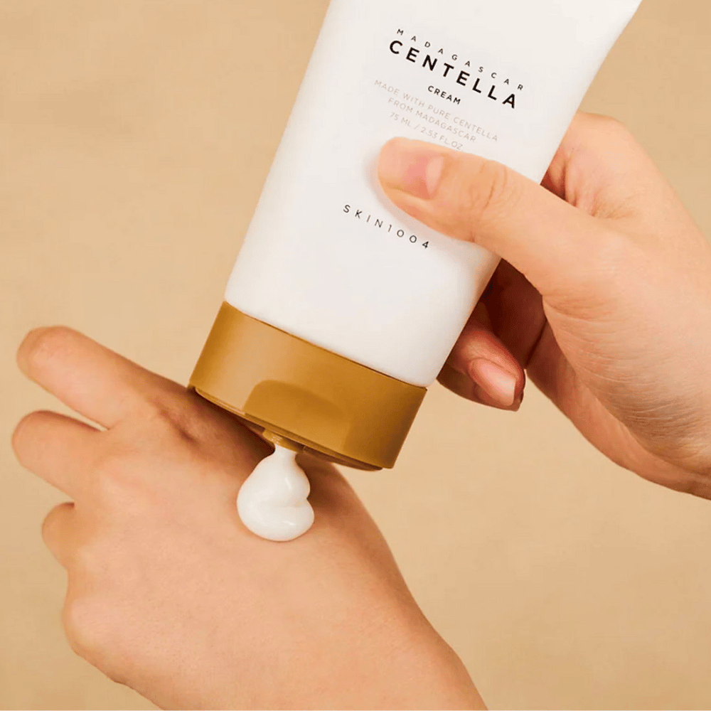 De afbeelding toont een persoon die een tube "Madagascar Centella Cream" van SKIN1004 vasthoudt. Het is een huidcrème die is gemaakt van pure Centella uit Madagaskar, in de grootte van 75 ml / 2.53 fl.oz. De crème wordt op de hand aangebracht en is bedoeld om de huid te voeden en te hydrateren.
