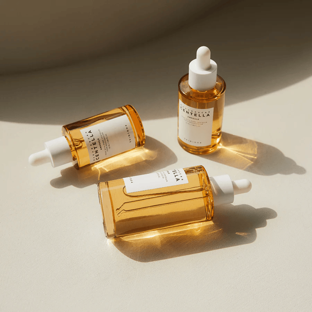 De afbeelding toont drie flessen van "Madagascar Centella Asiatica 100 Ampoule". Ze zijn geplaatst op een oppervlak waar zonlicht een prachtige reflectie creëert door de gouden inhoud van de flessen. Het product lijkt deel uit te maken van een huidverzorgingslijn die centella asiatica gebruikt, een ingrediënt dat bekend staat om zijn kalmerende eigenschappen. Het ontwerp van de flessen is eenvoudig en modern, wat suggereert dat het een premium huidverzorgingslijn is.