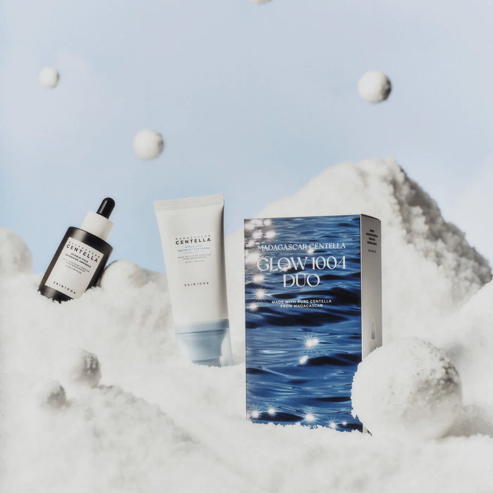 De afbeelding toont de huidverzorgingsset "Madagascar Centella Glow 1004 Duo" van SKIN1004, geplaatst tegen een achtergrond die sneeuw en hemel voorstelt.