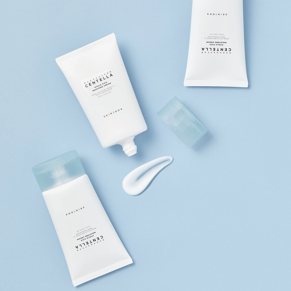 Twee witte tubes met SKIN1004 Centella huidverzorgingsproducten, een dotje product en een heldere blauwe dop, op een lichtblauwe achtergrond.