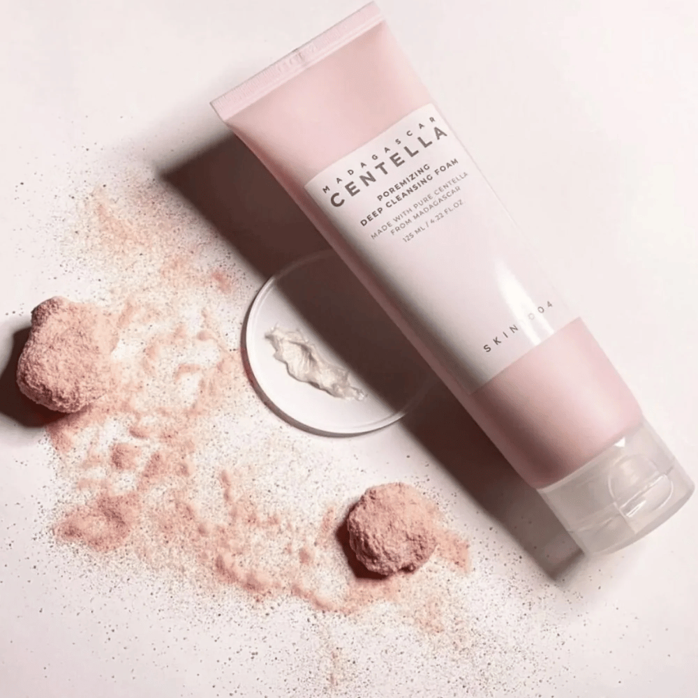 Madagascar Centella Poremizing Deep Cleansing Foam wordt gepresenteerd in een roze tube. De tubes liggen op een lichte ondergrond samen met roze poeder dat een gevoel van reinheid en frisheid geeft. Het product is ontworpen om de huid diep te reinigen met behulp van pure centella uit Madagaskar.