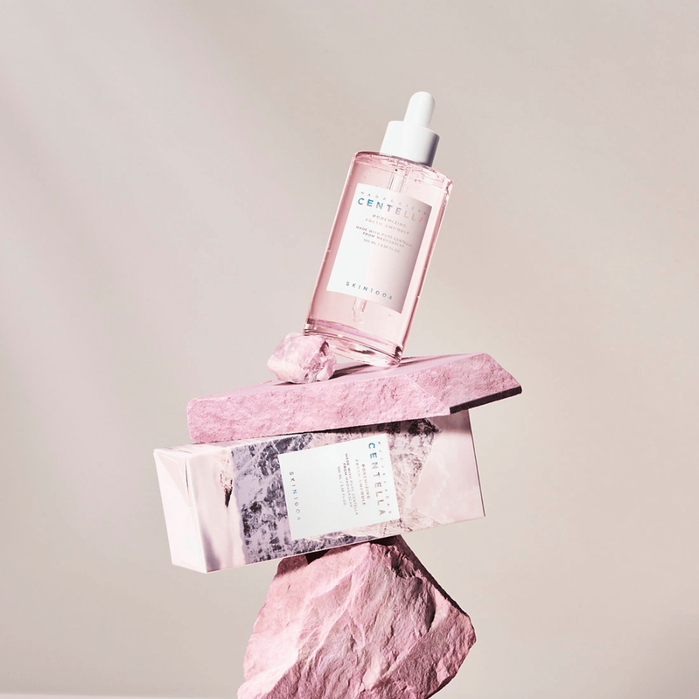 Doorzichtige fles met roze Centella-ampul van SKIN1004, gebalanceerd op stenen blokken, met een kartonnen verpakking eronder, tegen een neutrale achtergrond.