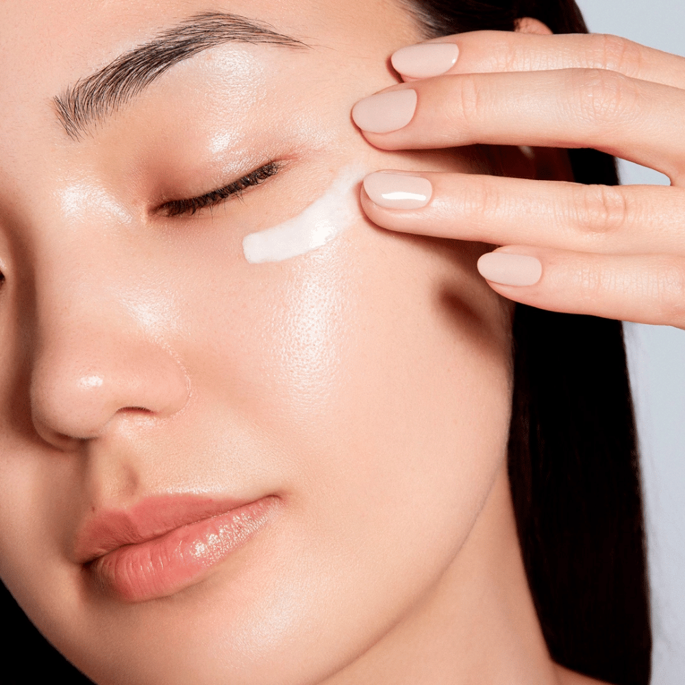 Een vrouw brengt voorzichtig een witte oogcrème aan onder haar oog. Haar huid ziet er stralend en gehydrateerd uit, wat het effect van de oogcrème benadrukt. Dit benadrukt het product als zowel hydraterend als geschikt voor de gevoelige huid rond de ogen.