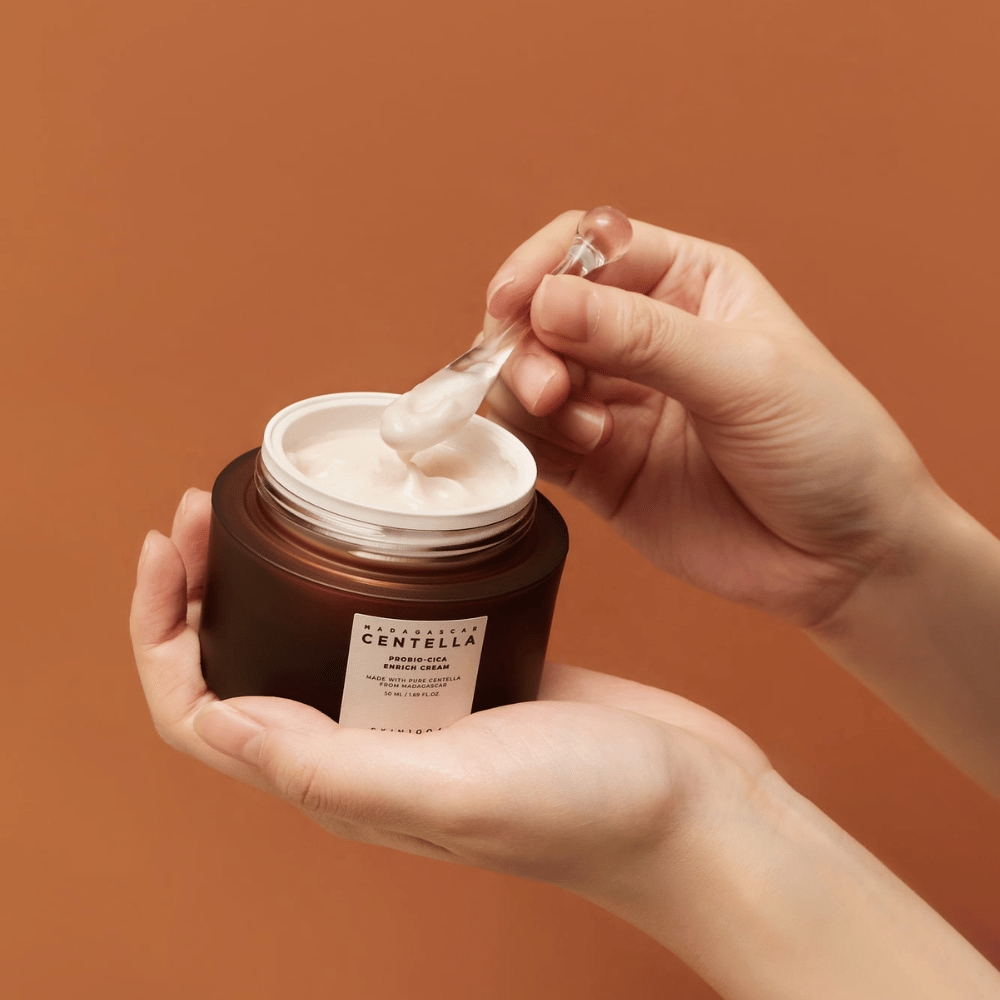 Handen die een open pot van Madagascar Centella Probio-Cica Enrich Cream vasthouden en een spatel met crème tegen een uniforme terracotta gekleurde achtergrond.