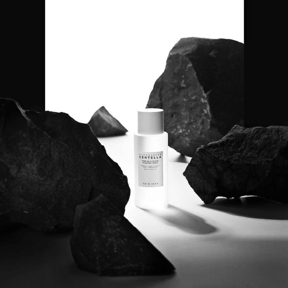 Witte fles SKIN1004 Madagascar Centella Tone Brightening Boosting Toner dramatisch geplaatst tussen grote zwarte stenen tegen een witte achtergrond.