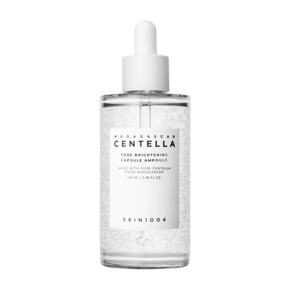 De afbeelding toont een fles van "SKIN1004 Madagascar Centella Tone Brightening Capsule Ampoule". Het is een doorzichtige fles met een witte druppelaar. Op het etiket staat dat het product pure centella-ingrediënten uit Madagaskar bevat. De inhoud van de fles lijkt kleine capsules of deeltjes te hebben, wat deel kan uitmaken van de verhelderende huidverzorgingseigenschappen. De fles bevat 100 ml / 3.38 fl.oz. van het product.