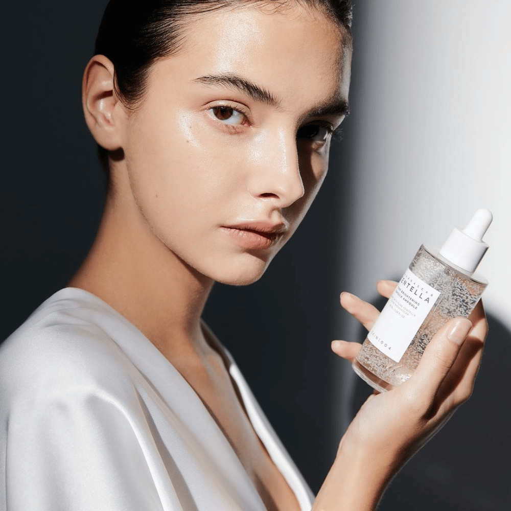 Op de afbeelding is een persoon te zien die een fles met een druppelaar vasthoudt. De fles is gemerkt "SKIN1004 Madagascar Centella Tone Brightening Capsule Ampoule". De persoon heeft een glanzende huidtint die het licht weerkaatst, wat wijst op een mogelijk effect van het product.