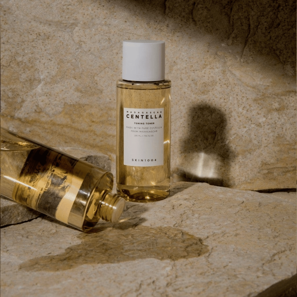 De afbeelding toont een fles "Madagascar Centella Toning Toner" van SKIN1004. Het is een doorzichtige fles gevuld met een gelige vloeistof, waarschijnlijk een toner voor huidverzorging. Er staat dat het gemaakt is met pure centella uit Madagaskar en de inhoud is 210 ml / 7.10 fl.oz. De fles en een liggende fles zijn geplaatst op een rotsachtige ondergrond, wat een natuurlijke en minimalistische uitstraling geeft.
