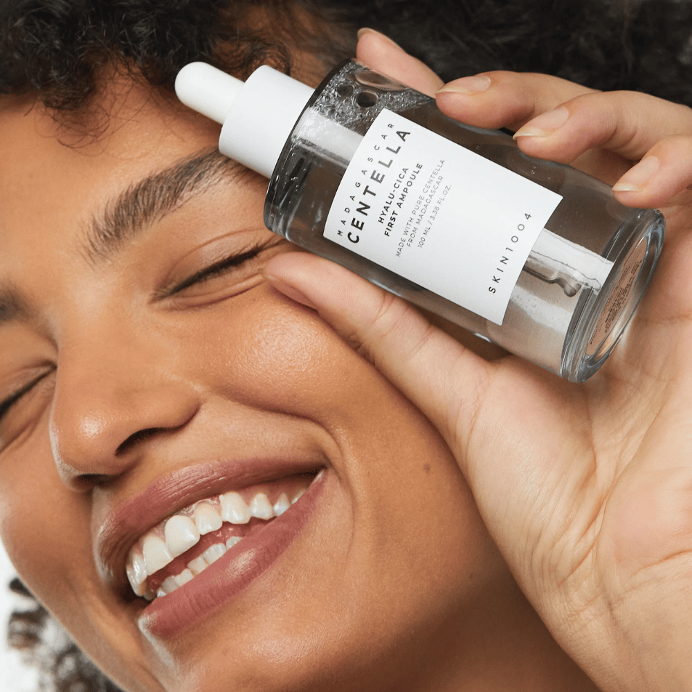 Lachende vrouw houdt Skin1004 Madagascar Centella Hyalu-Cica First Ampoule naast haar gezicht omhoog.