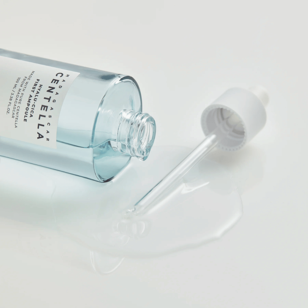 Close-up van een open fles Skin1004 Madagascar Centella Hyalu-Cica First Ampoule met liggende pipet en serum verspreid op een wit oppervlak.