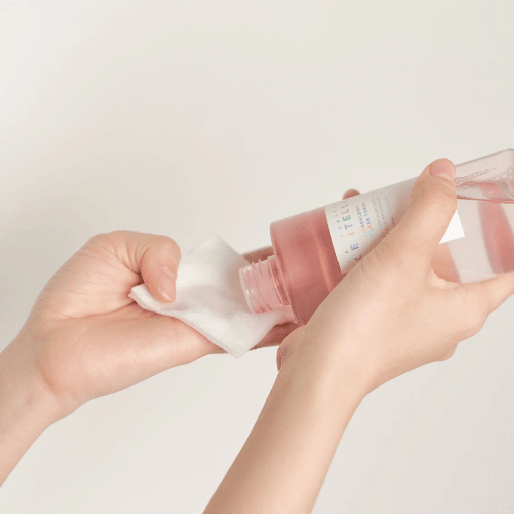 Een persoon giet Madagascar Centella Poremizing Clear Toner uit een transparante fles op een wattenschijfje. Het product, dat een roze tint heeft, wordt gebruikt om de huid op te frissen en te reinigen. De handen zijn zichtbaar, wat het aanbrengen van de toner laat zien.
