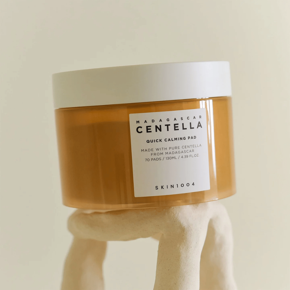 Pot met SKIN1004 Madagascar Centella Quick Calming Pad geplaatst op een sculptuurachtige witte sokkel – amberkleurige container met wit etiket en deksel.