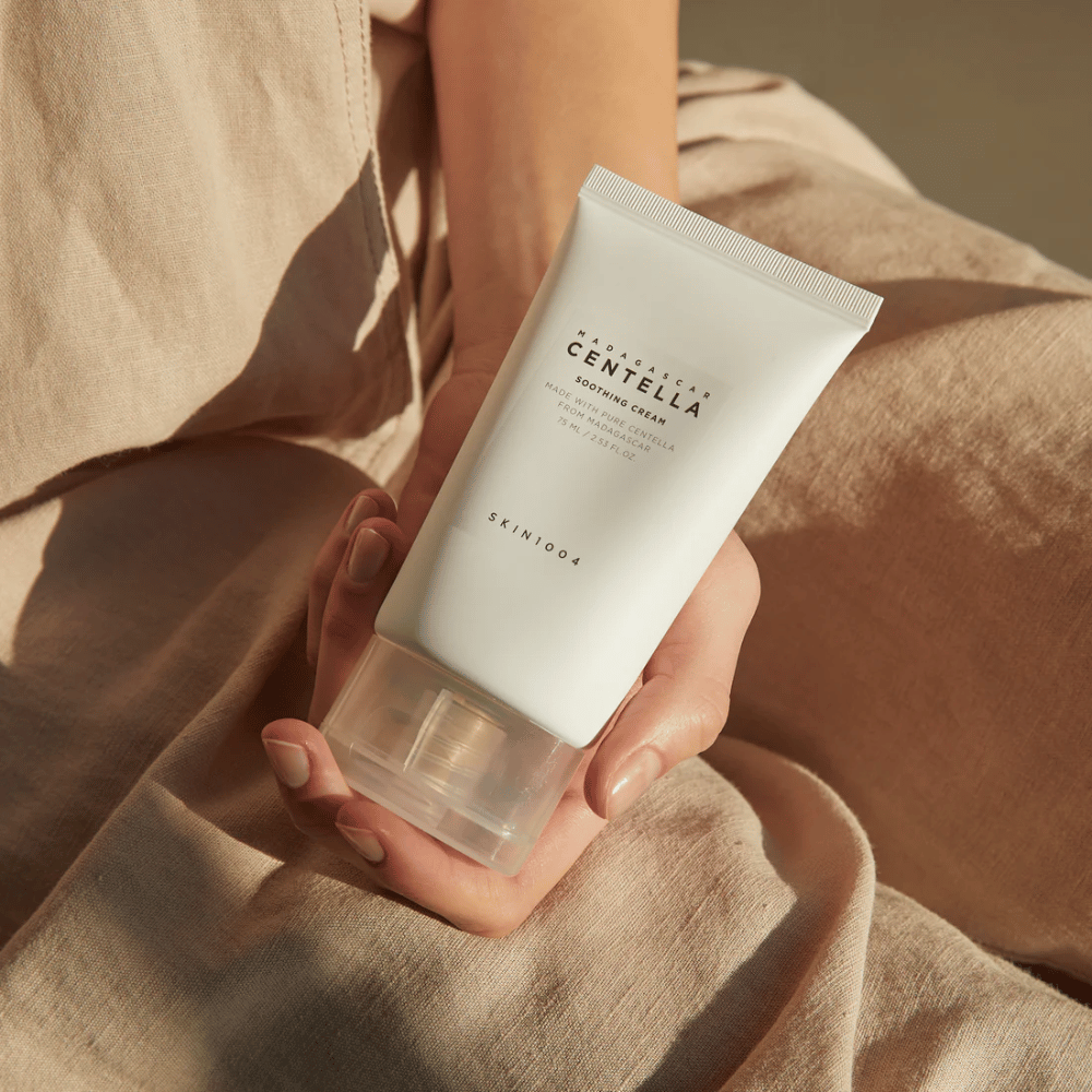 Een persoon houdt een witte tube van Madagascar Centella Soothing Cream van SKIN1004 vast. De tube heeft een doorzichtige dop en tekst die het product beschrijft als kalmerend en gemaakt met pure Centella. De persoon is gekleed in lichte kleding.