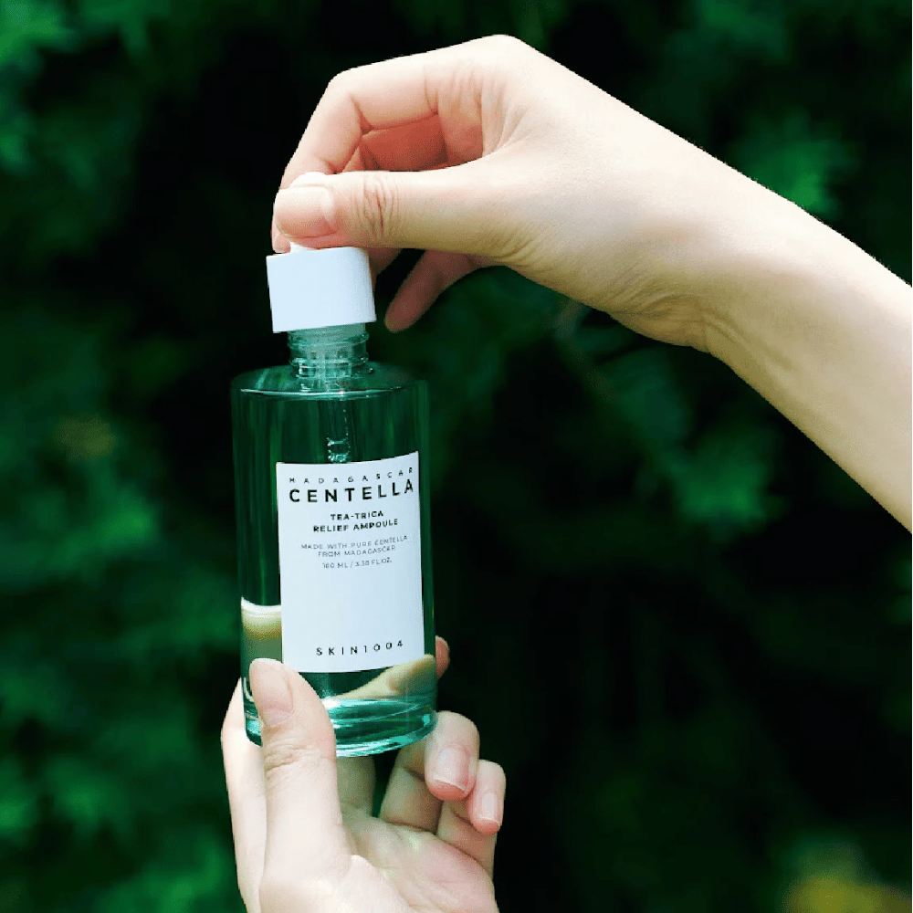 Persoon opent SKIN1004 Madagascar Centella Tea-Trica Relief Ampoule – groene glazen fles met wit etiket wordt in handen gehouden voor groene achtergrond.