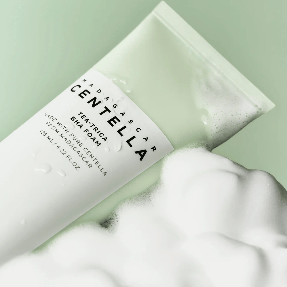 Een product van het type reinigingssop, "Madagascar Centella Tea-trica BHA Foam", is gedeeltelijk zichtbaar met schuim eromheen. Het product heeft een groen en wit ontwerp, en de tekst "Madagascar Centella" en "Tea-trica BHA Foam" zijn duidelijk zichtbaar. Het volume is 125 ml.