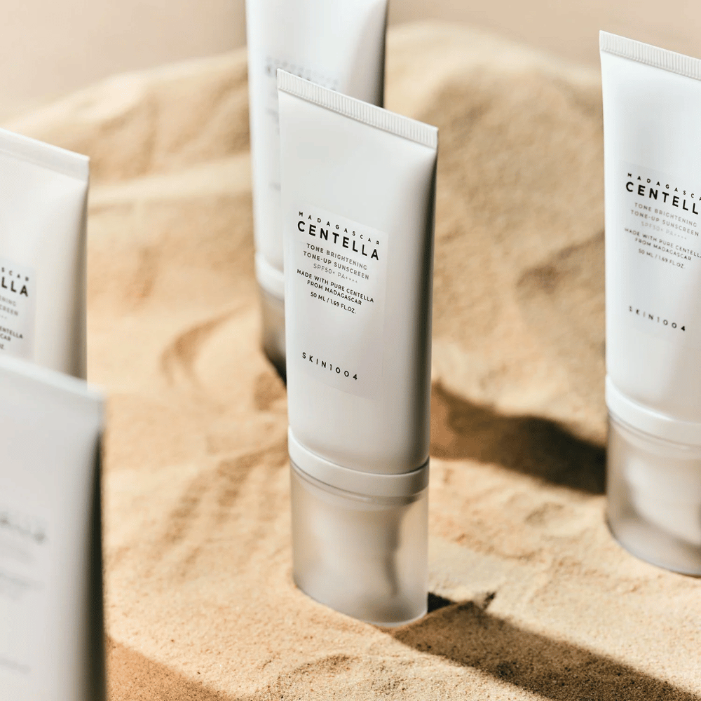 Meerdere tubes met Madagascar Centella Tone Brightening Tone-Up Sunscreen staan in het zand. De tube is wit met zwarte tekst en bevat 50 ml. Het product heeft SPF 50+ PA++++ en is gemaakt met zuivere Centella uit Madagaskar.