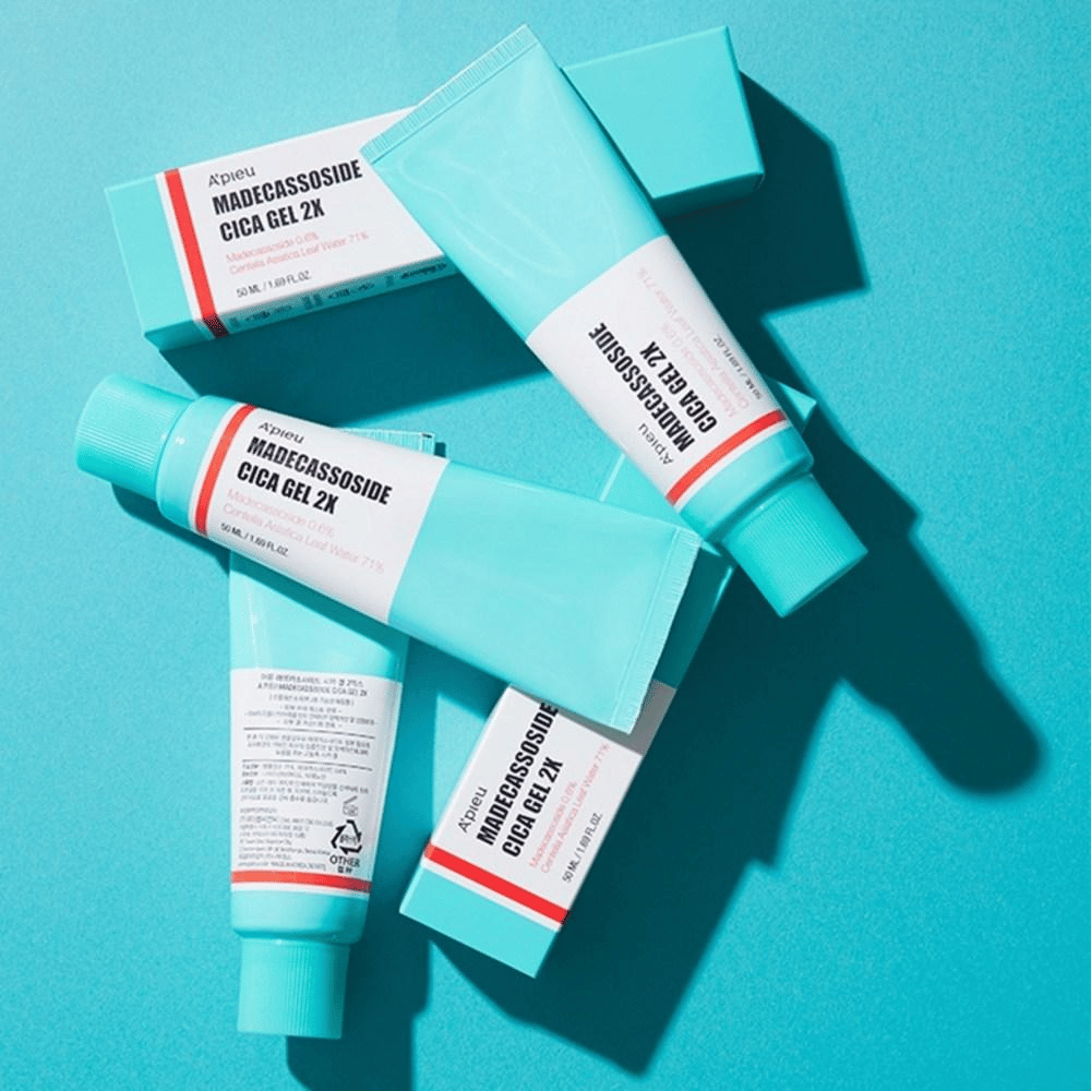 Meerdere tubes en dozen van A’pieu Madecassoside Cica Gel 2X in een turquoise verpakking met wit en rood etiket, liggend tegen een turquoise achtergrond in fel licht.
