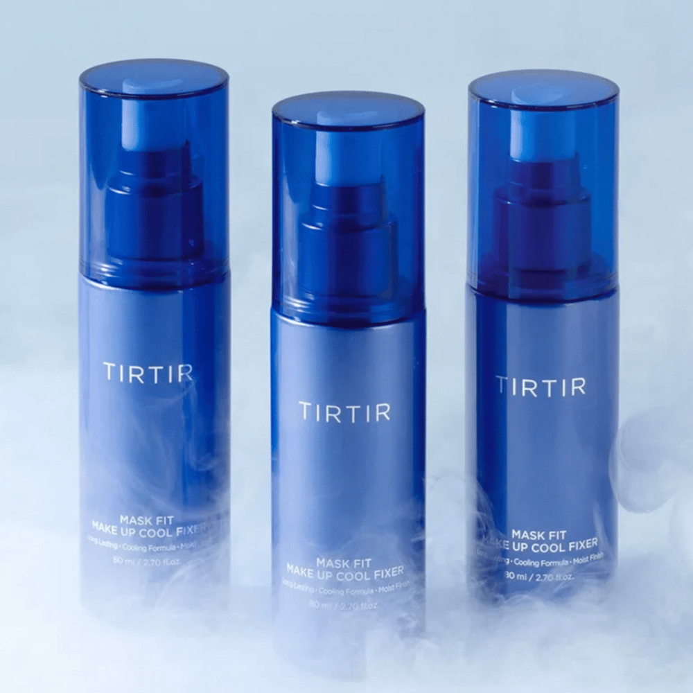 Drie blauwe spuitflessen met "TIRTIR Mask Fit Make Up Cool Fixer" op het etiket. Elke fles bevat 80 ml (2,70 fl. oz.) en is ontworpen om een verfrissend gevoel en langdurige make-up te bieden. De flessen staan op een lichte achtergrond met mist om een sfeer van frisheid te creëren.