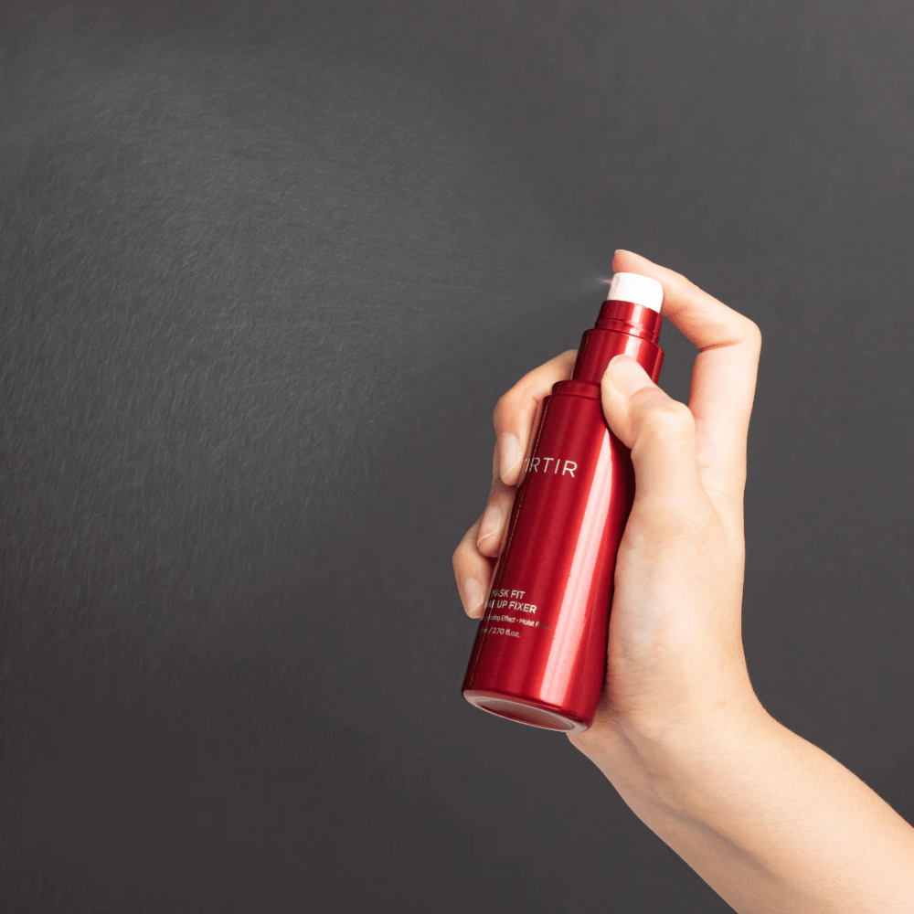 Een hand die een rode fles van het merk tirtir Mask Fit Make Up Fixer vasthoudt en een fijne mist sprayt, ideaal voor het fixeren van make-up.