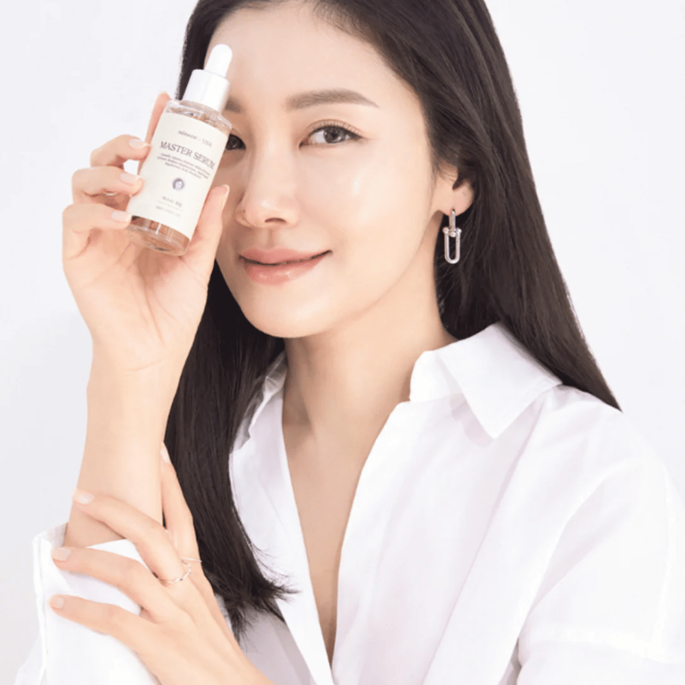 Glimlachende vrouw met lang, donker haar en een witte blouse houdt Mixsoon Master Serum naast haar gezicht. Haar huid ziet er stralend en gehydrateerd uit.