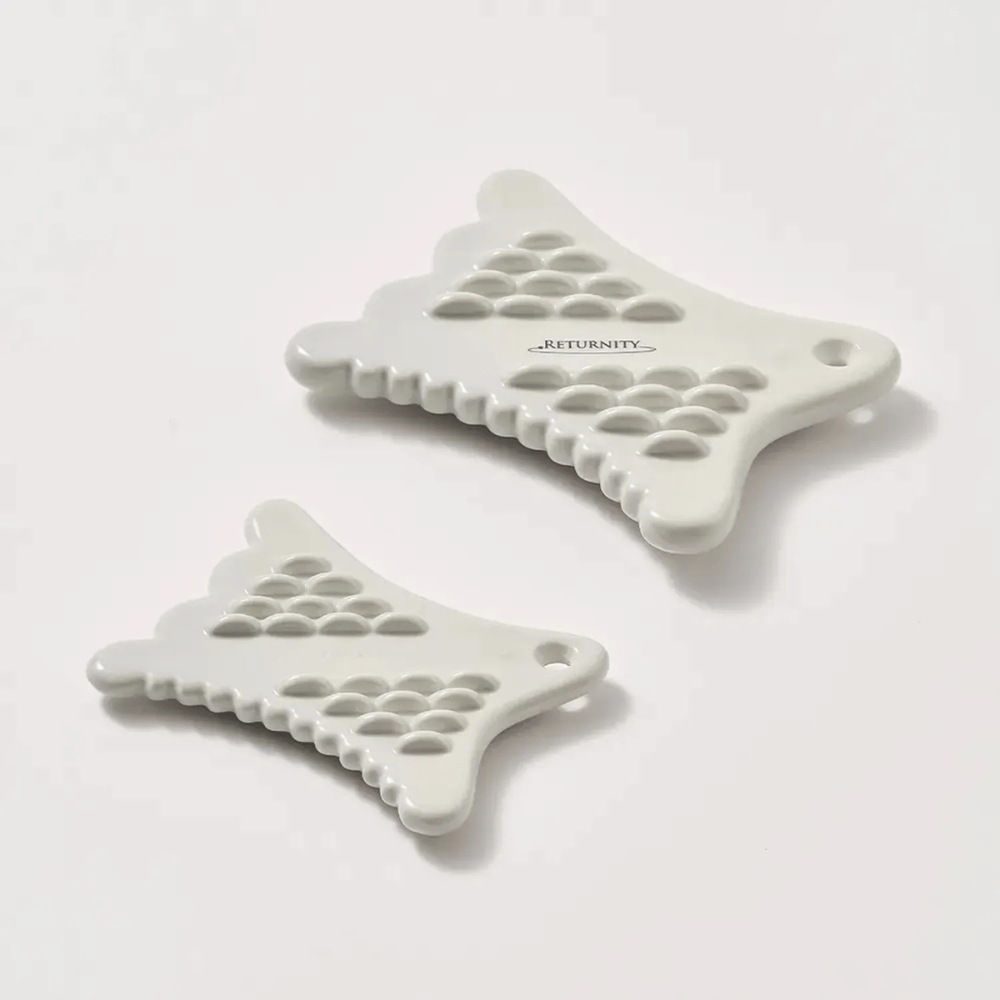 Twee witte RETURNITY Mini Guasha-tools met gestructureerd oppervlak en ergonomisch ontwerp op een lichte achtergrond.