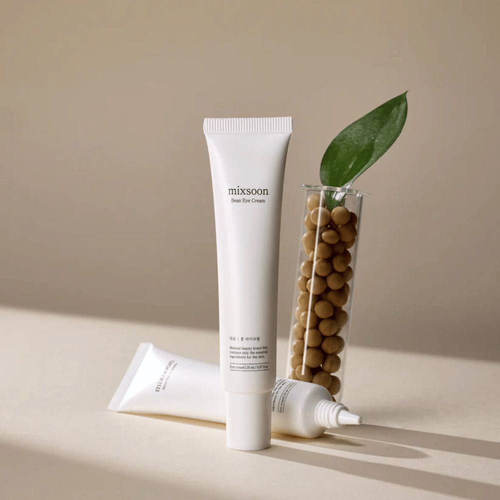 Twee tubes Mixsoon Bean Eye Cream geplaatst naast een reageerbuis gevuld met sojabonen en een groen plantblad, op een beige oppervlak met schaduwspel.