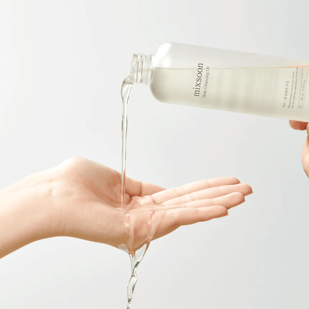 Een heldere reinigingsolie wordt uit een transparante fles met wit etiket waarop "MixSoon" staat, in een open handpalm gegoten. De olie vormt een glanzende straal tussen de opening van de fles en de hand, waarmee de vloeibare consistentie van de olie wordt gedemonstreerd. De hand is stil en wordt zo gehouden dat het de hele hoeveelheid reinigingsolie kan opvangen tegen een effen lichte achtergrond.