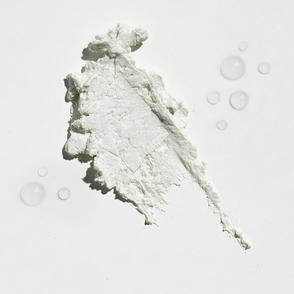 Close-up van een witte, romige substantie op een witte achtergrond, met enkele waterdruppels eromheen. De textuur lijkt op een zonnepoeder of foundation met een lichte gloed.