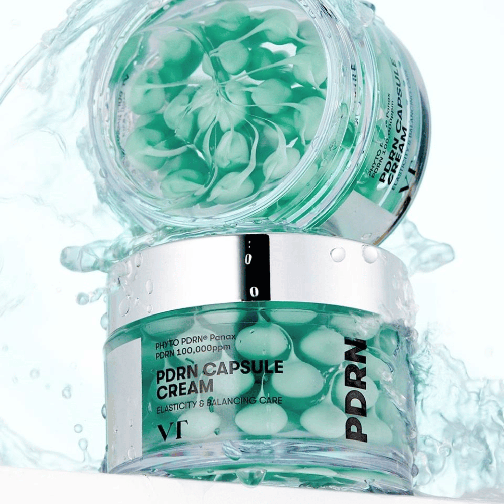 Twee potten van VT PDRN Capsule Cream gestapeld met waterspatten eromheen. De doorzichtige potten tonen groene capsules binnenin, met de tekst "Elasticity & Balancing Care" zichtbaar op het etiket.