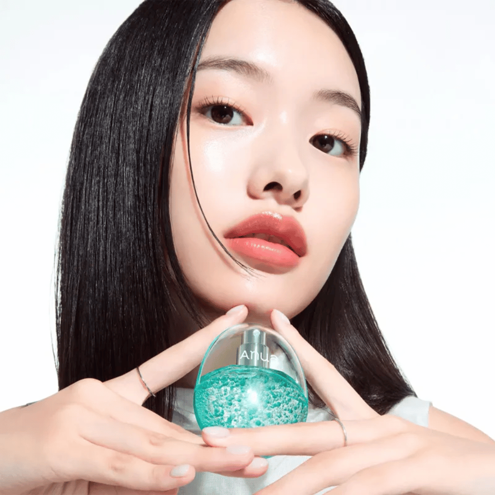 Close-up van een model dat een ronde, transparante Anua-fles vasthoudt, gevuld met turquoise capsules of gelparels. De afbeelding legt de focus op de glans van de huid en het innovatieve design van het product, wat een frisse en moderne indruk geeft.
