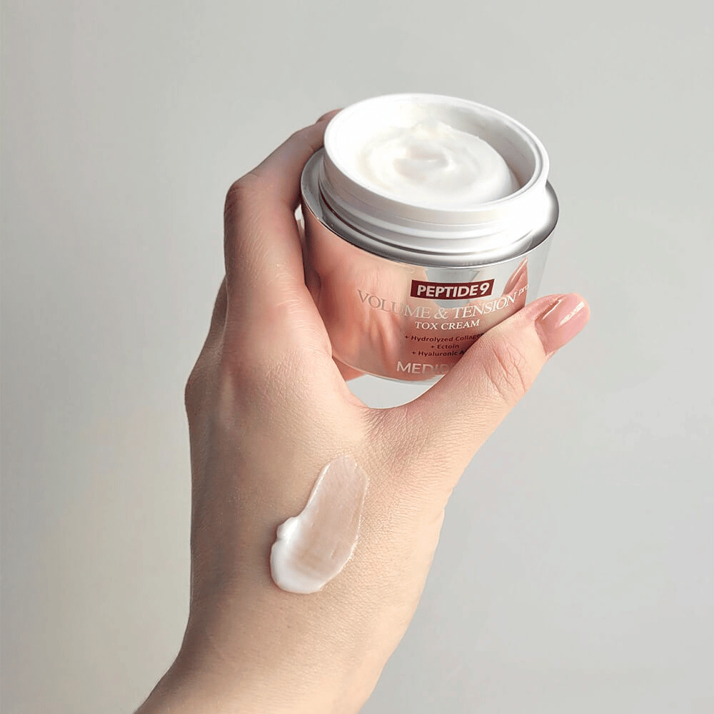 Hand houdt MEDI-PEEL Peptide 9 Volume & Tension Tox Cream vast met een druppel crème op de rug van de hand. De zijdezachte formule hydrateert en geeft de huid stevigheid en veerkracht.