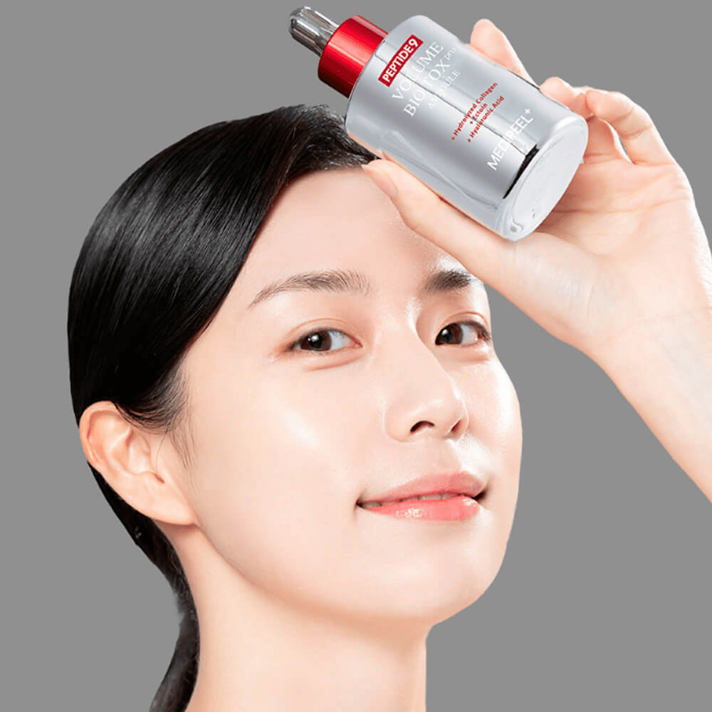 Persoon houdt MEDI-PEEL Peptide 9 Volume Bio Tox Pro Ampoule tegen het voorhoofd. De huid ziet er gehydrateerd en stralend uit - het serum versterkt elasticiteit en stevigheid.